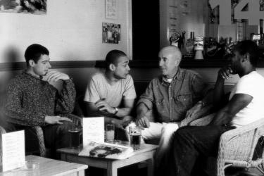 Four men sitting talking around a coffee table in a posed photograph to promote the Metro Thrust HIV prevention and sexual health outreach project, 1995