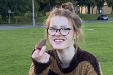 Smiling, caucasian teenager. Her hair is up and she is wearing glasses and a baggy brown jumper, holding a chocolate bar.