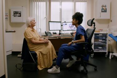 Wide angle photo from a healthcare setting. A white haired white woman sits, having a consultation with a young mixed race woman wearing hospital scrubs