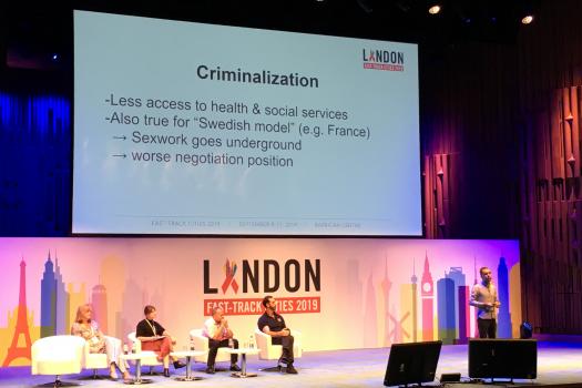 Slide on Criminalisation above panel and speaker on stage at London Fast-Track Cities Conference