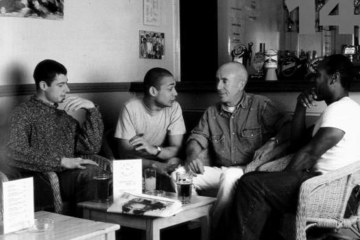 Four men sitting talking around a coffee table in a posed photograph to promote the Metro Thrust HIV prevention and sexual health outreach project, 1995