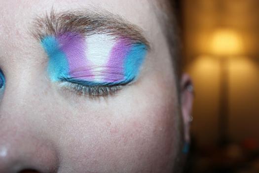 Photo cropped around a young person's eye, which is closed with the colours of the trans flag on their eyelid