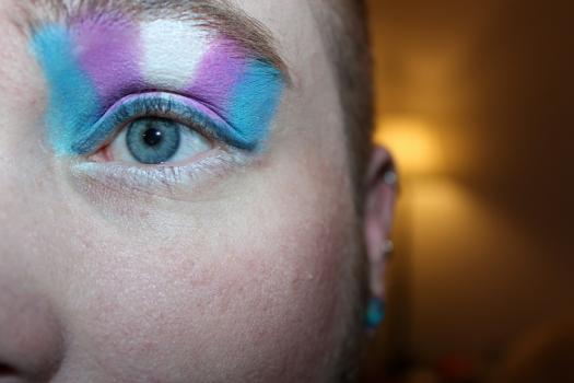 Photo of a person's face cropped around their open eye with trans flag eye shadow