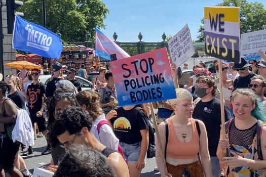 Photo of people at Trans Pride London, holding placards: 'Stop policing bodies' and 'We exist' on a Asexual flag background