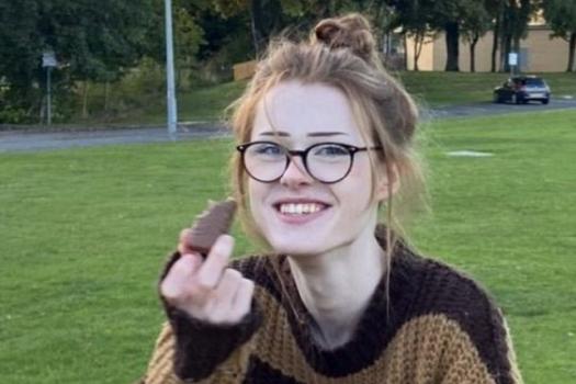 Smiling, caucasian teenager. Her hair is up and she is wearing glasses and a baggy brown jumper, holding a chocolate bar.