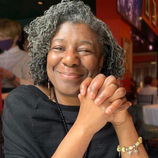 Photo of Debbie Solomon, smiling at the camera. She is a Black woman with chin-length salt-and-pepper locks. She is sitting at a restaurant with red walls.