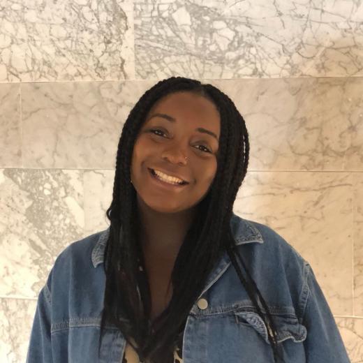 Photo of Trustee Joanna Webley-Brown, a young Black woman with box braids. She is smiling at the camera, stood in front of a pale wall