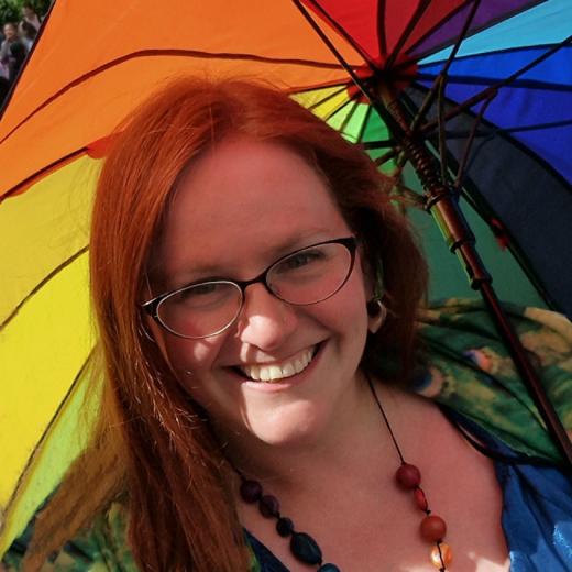 Caucasian woman with long red hair and glasses smiling at the camera. She is holding a rainbow umbrella